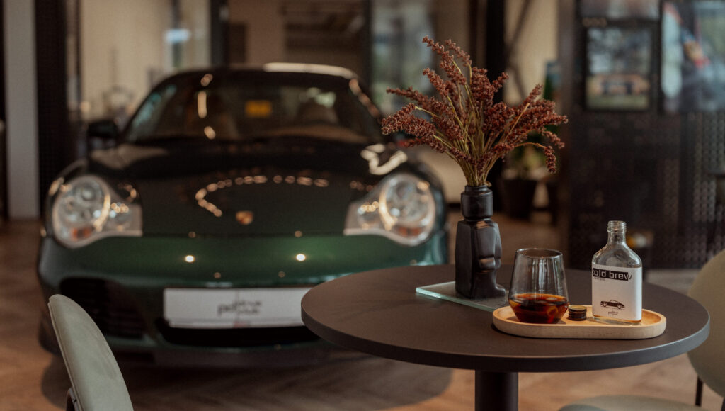 Pohár a fľaša cold brew na okrúhlom stole, v pozadí zelené Porsche 911 (996) v showroome.