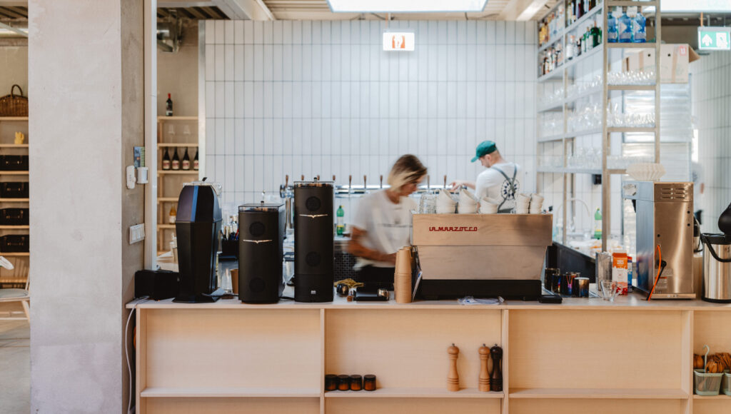 Bar Ferdinand: nerezový kávovar La Marzocco, dva čierne mlynčeky Victoria Arduino, biely obklad, otvorené regály s pohármi, baristi v pohybe.