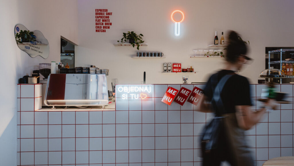 Interiér kaviarne Jovipoint s bielo-červeným obkladom a neónmi; na bare pákový kávovar Nuova Simonelli Eagle One; barista v pohybe.