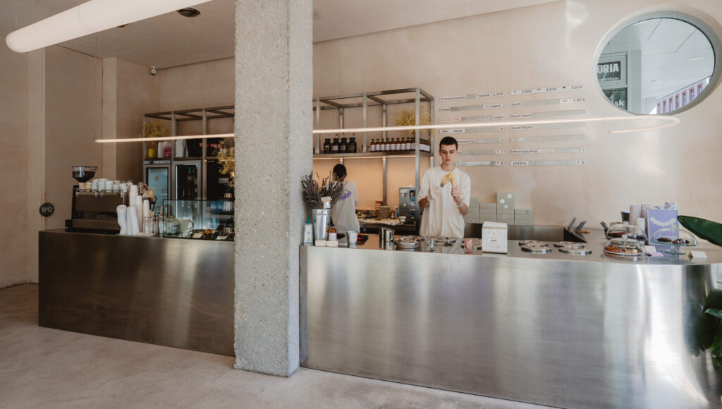 Interiér Mimóza: barista naberá zmrzlinu za nerezovým pultom v minimalistickej kaviarni s menu na stene.