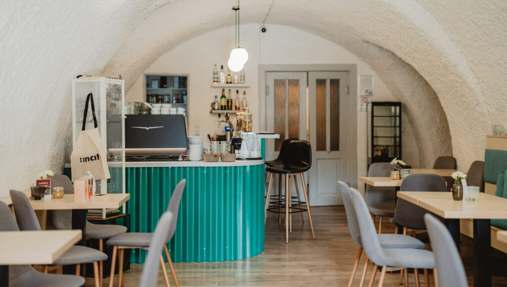 Interiér kaviarne Incite: biely klenbový strop, zelený oblý bar s profesionálnym espresso kávovarom Victoria Arduino, sivé čalúnené stoličky a svetlé stoly.