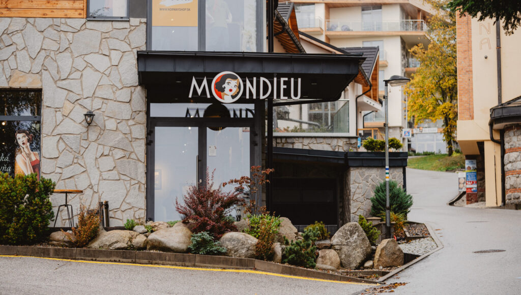 Vstup do Mondieu s čiernou markízou a logom, kamenná fasáda, zeleň a úzka ulica v pozadí.