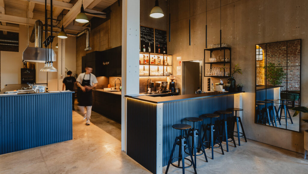 Minimalistický bar s rebrovaným modrým pultom, vysokými stoličkami a tlmeným závesným osvetlením v industriálnom priestore; v pozadí otvorená kuchyňa a poličky s pohármi.