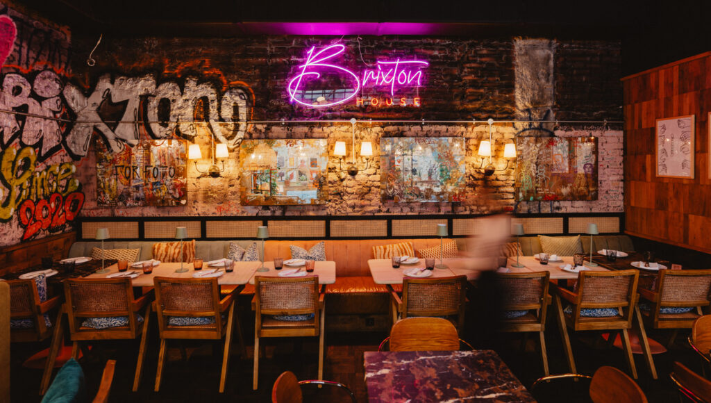 Interiér reštaurácie Brixton House: tehlová stena s graffiti a fialovým neónom, nástenné lampy a prestreté drevené stoly pre skupiny.
