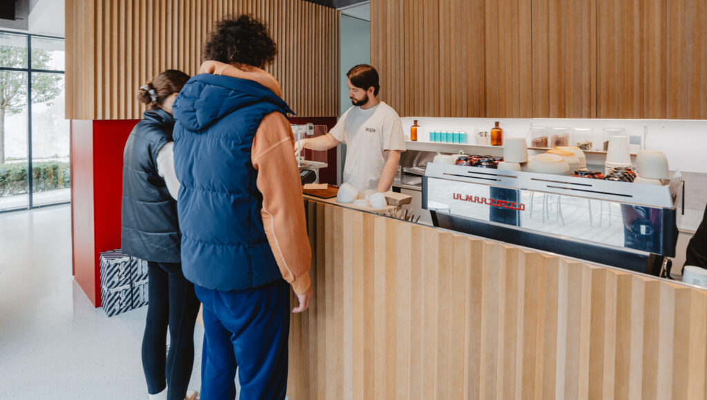 Barista pri pulte pripravuje kávu na kávovare La Marzocco v modernej kaviarni Mandľa s dreveným interiérom a zákazníkmi.