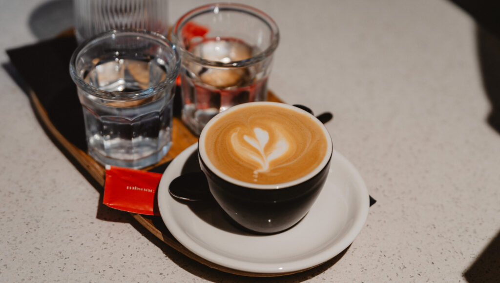Cappuccino s tulipánovým latte artom v čiernej šálke, na drevenej tácke s dvomi pohármi vody – Rebarbora.
