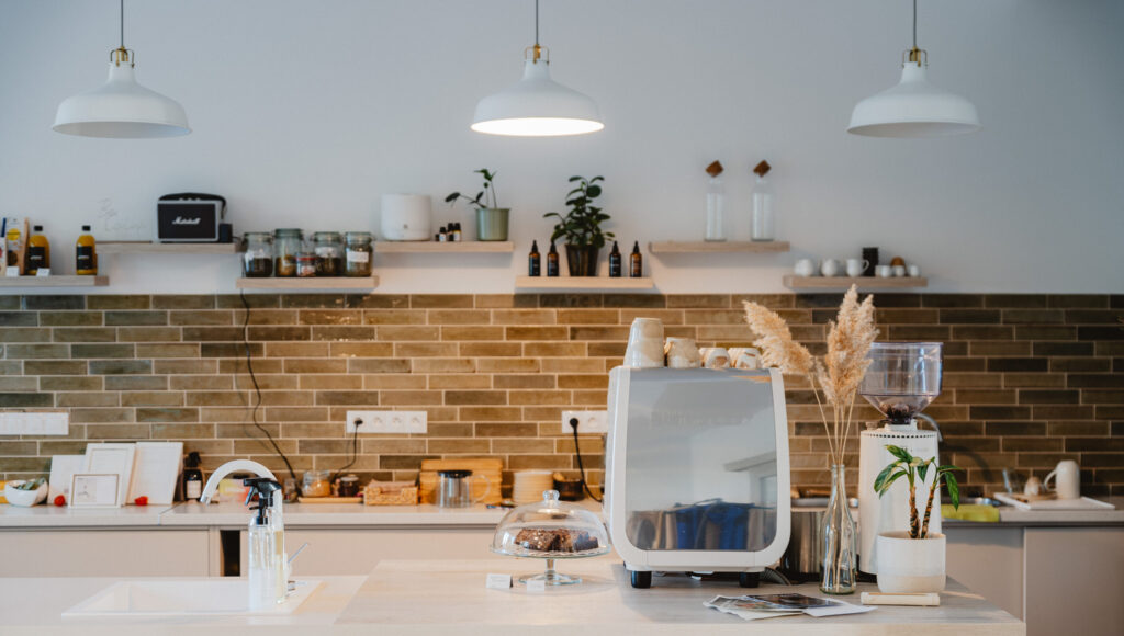 Svetlý kávový kútik Calma: biely kávovar Nuova Simonelli, mlynček Nuova Simonelli, sklenený stojan s brownies, pampová tráva vo váze, zelený tehlový obklad a poličky s rastlinami a dózami.