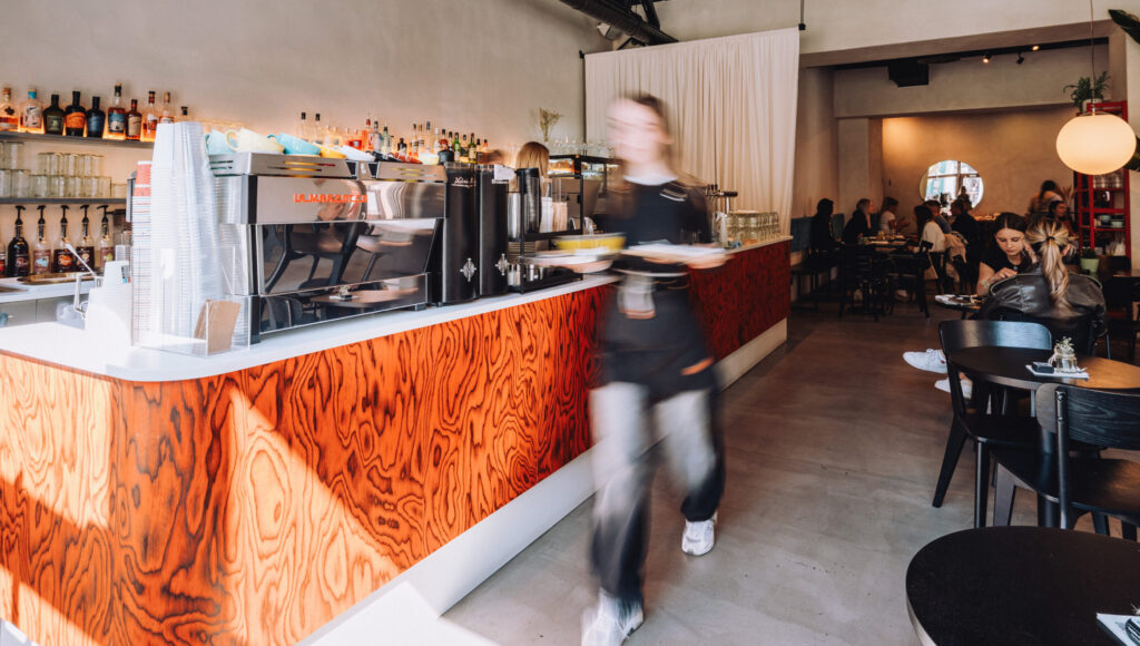 Interiér kaviarne Paddock – výrazný drevený bar, kávovar La Marzocco, čašníčka s taniermi a hostia v pozadí.